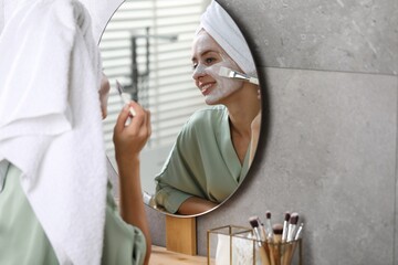 Woman applying face mask in front of mirror indoors. Spa treatments