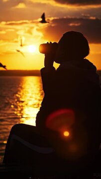 Vertical silhouette young woman drinks coffee by orange sunset sky near water in slow motion. A girl in setting sun rays sits on a parapet with coffee in her hands and enjoys life. Healthy lifestyle