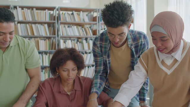 Tilt shot of excited young multiracial migrant students playing card game with English verbs while practicing grammar together during interactive lesson in library