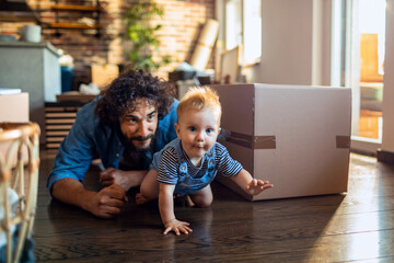 A playful father and his curious baby explore the floor near a moving box in their cozy home