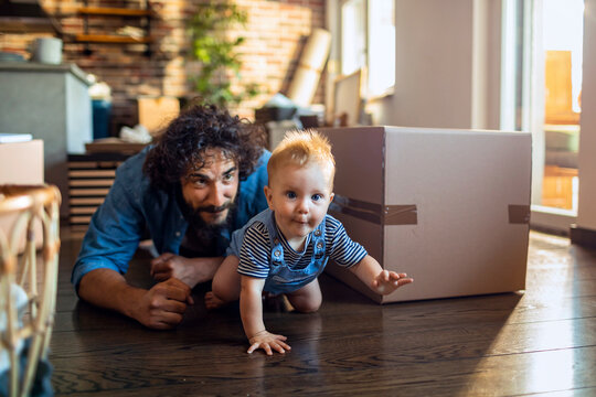 A playful father and his curious baby explore the floor near a moving box in their cozy home