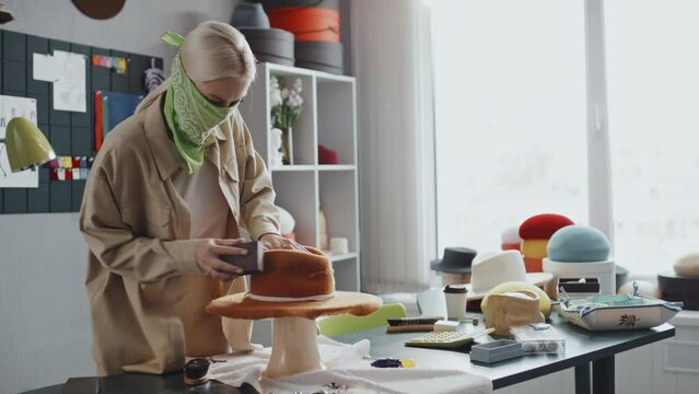 Medium shot of young female hat-maker wearing green cloth aroundher face for protection while cleaning new felt hat with special brush