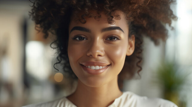 Smiling young woman with curly hair, possibly of African descent, in a casual, plant-filled environment exuding bright cheerfulness