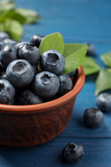 Tasty fresh blueberries with leaves in bowl on blue wooden table, closeup