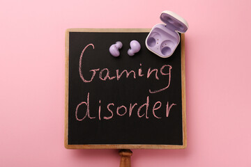 Small chalkboard with written phrase Gaming Disorder and earphones on pink background, top view