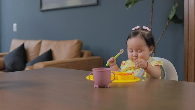 Baby eating with his hands in the dining room at home and holding a spoon in his other hand, baby led weaning concept