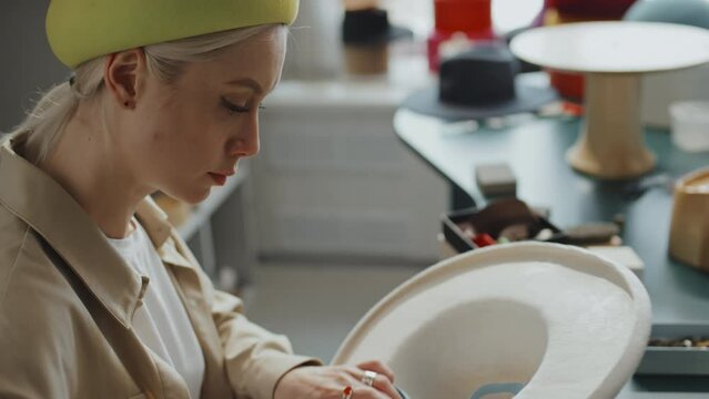 High angle shot of young Caucasian woman sitting in her studio pleating blue hat lining into white cowboy hat