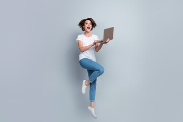 Full size photo of impressed crazy girl dressed white t-shirt jeans jump hold laptop read notification isolated on gray color background
