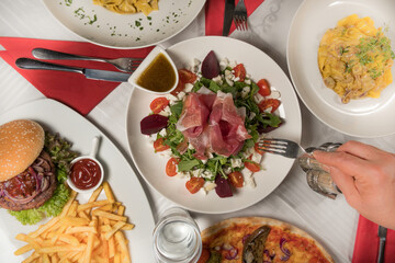 A plate of arugula lettuce with ham and tomatoes surrounded by a hamburger with onions and  fries, pizza and pasta.