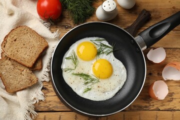 Frying pan with tasty cooked eggs, dill and other products on wooden table, flat lay