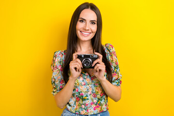 Photo of toothy beaming cute girl with long hair dressed flower print blouse arms holding camera isolated on yellow color background - Powered by Adobe