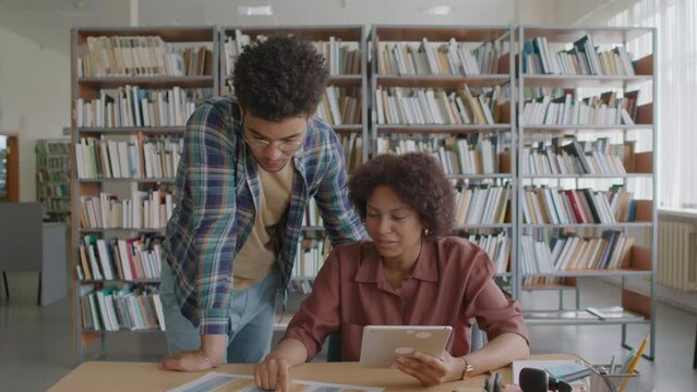Medium shot of two multiracial migrant students using digital tablet and list of irregular English verbs while doing home assignment together in modern library