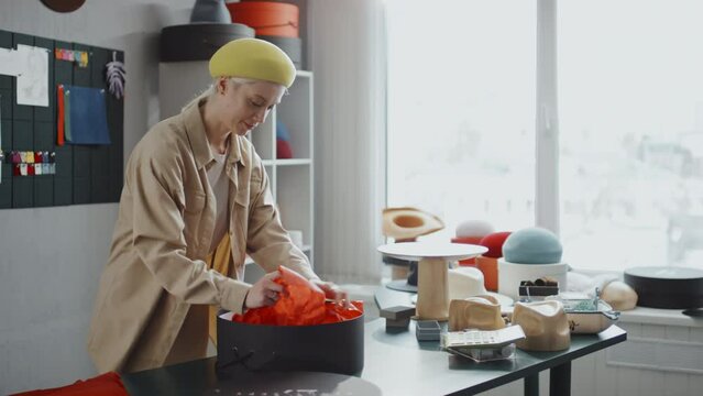 Medium full shot of Caucasian female hat-maker packaging her creation using red paper and big black gift wrap