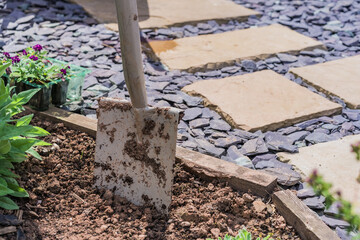 A close-up view of a dirty garden spade embedded in freshly turned soil, next to potted purple flowers and lush green plants.