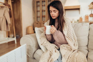 Young woman in warm clothes and plaid holding cup of hot drink while sitting on couch at home near...