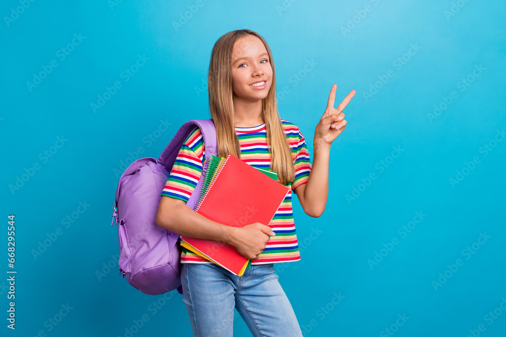 Poster Photo of positive schoolgirl dressed striped t-shirt rucksack on shoulder hold copybook show v-sign isolated on blue color background