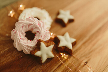 New Year background for winter cards. Gingerbread cookies in the shape of stars and pink meringue cookies lie on a brown background with Christmas lights. Delicious sweet pastries on a wood table