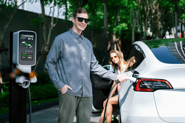Young couple travel with EV electric car charging in green sustainable city outdoor garden in...