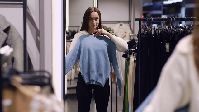 An adult girl tries on a blue sweater while looking in the mirror in a store. A young woman chooses clothes in a store while shopping. Seasonal holiday discounts on purchases. Black Friday and Sale