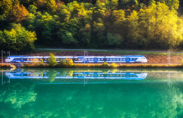 Beautiful blue modern high speed train and river in alpine mountains at sunrise in autumn. Passenger train, reflection in water, railroad, lake, orange trees in fall. Railway station in Slovenia