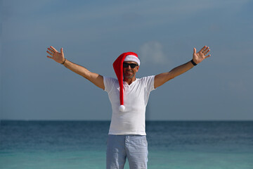 A tanned man in a Santa Claus hat and sunglasses, shorts and a white T-shirt stands on a snow-white...