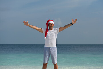 A tanned man in a Santa Claus hat, a white T-shirt, blue shorts, stands on a snow-white tropical...