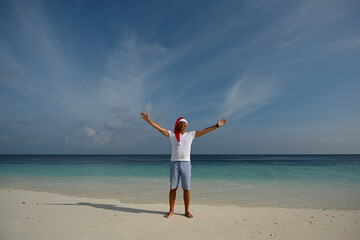 A tanned man in a Santa Claus hat, a white T-shirt, blue shorts, stands on a snow-white tropical...