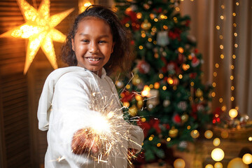 Cheerful dark-skinned teenage girl holding sparklers, near the Christmas tree at home. Christmas...
