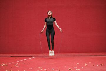 Sporty young girl in sportswear conducts a workout. Female athlete jumps on a rope against the background of a red wall in the park.