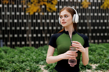 Sporty young girl in sportswear conducts a workout. The athlete is resting in headphones and drinking water in the autumn park.