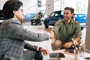 Selective focus of cheerful customer male purchasing automobile in dealership signing paper shaking hands with dealer enjoying successful agreement. Concept of buying new auto at showroom.