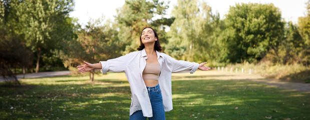 Carefree asian girl laughing and dancing in park, enjoying summer sunny day, raising hands up and breathing fresh air