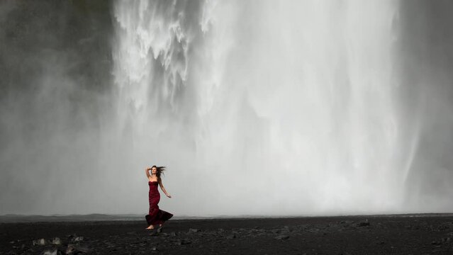 Breathtaking view of woman overwhelmed with happieness on the background of huge amount of spray the waterfall produces, falling on black-sanded beach. High quality 4k footage