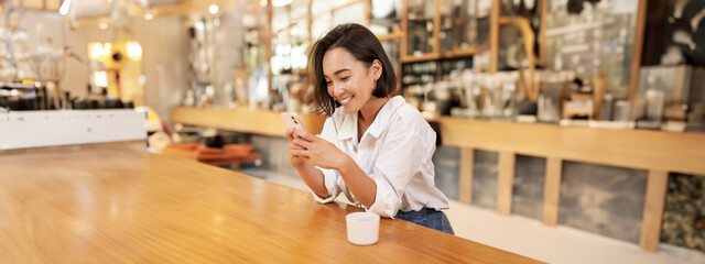 Vertical portrait of stylish asian woman sitting in cafe, drinking coffee and using smartphone