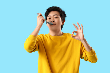 Young Asian man with spoon showing OK on blue background - obrazy, fototapety, plakaty