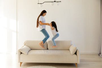 Happy mom and little daughter holding hands jumping on sofa together, baby sitter or mother...