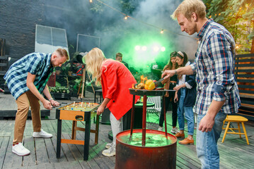 Handsome man grilling vegetables, other friends talking together near dj and one guy and girl play table football game. © gorynvd