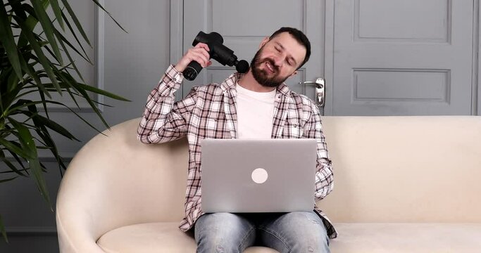 Massage gun muscle recovery therapy. Back pain stimulation. Bearded man working on laptop and using percussion massage gun. 