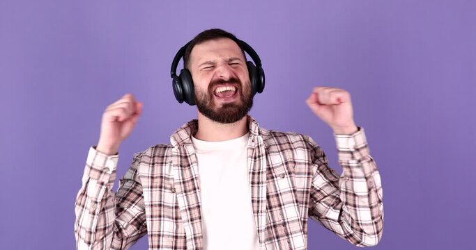 Portrait of attractive laughing bearded man wearing large wireless headphones on purple background.