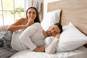 Awake young woman showing silence gesture with her sleeping husband in bedroom