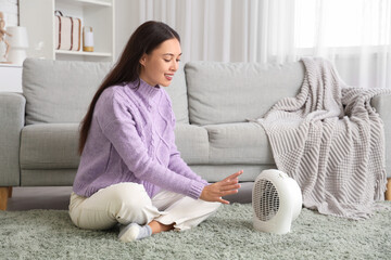 Pretty young woman warming hands near electric fan heater in living room