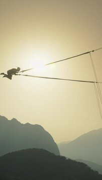 Vertical video. The Black Silhouette of an Extreme and Courageous Man against the Backdrop of the Sun Setting behind the Mountains. He is Attached to a Bungee Cord and Spins around at a Tremendous Hei