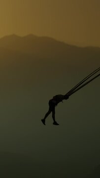 Vertical video. A Daring Man Who Loves Thrills and Adrenaline, He's Attached to a Bungee Cord in the Mountains and Gets Spun High in the Air. He Experiences Immense Pleasure and Moves His Legs as If W