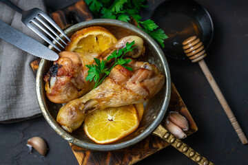 Chicken legs cooked in the oven in honey sauce with oranges, in a frying pan, on a board and on a dark table with a napkin, parsley and honey.