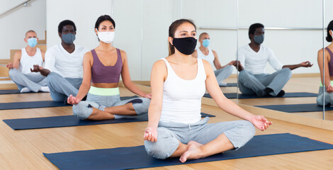 Multiethnic group of young adults in face masks for viral protection sitting in lotus position practicing meditation at yoga class