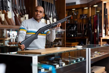 Rolgordijnen Hispanic man owner of armory shop offering modern hunting pump action shotgun. © JackF