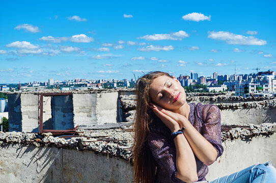 
A girl who is not afraid of heights, with a calm face, sits on the precipice of a cliff against the backdrop of the city and the blue cloudy sky.
