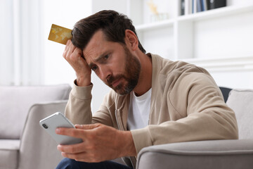 Upset man with credit card using smartphone in armchair at home. Be careful - fraud