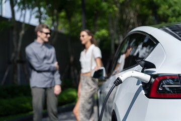 Fototapeta na wymiar Young couple travel with EV electric car charging in green sustainable city outdoor garden in summer shows urban sustainability lifestyle by green clean rechargeable energy of electric vehicle innards