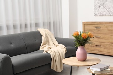 Vase with beautiful flowers and books on wooden nesting tables near grey sofa in room